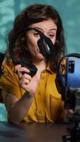 Vertical Audio engineer recording for internet channel in dimly lit home studio, presenting pairs of gaming headphones to audience. Woman showing viewers music listening device, camera A video