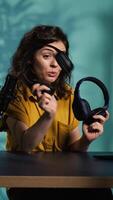 Vertical Audio engineer recording for internet channel in dimly lit home studio, presenting pairs of gaming headphones to audience. Woman showing viewers music listening device, camera B video