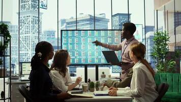Business associates collaborating at the conference table in boardroom, utilizing smart technology and monthly statistics to present analysis of market trends and economic forecasts. Camera B. video