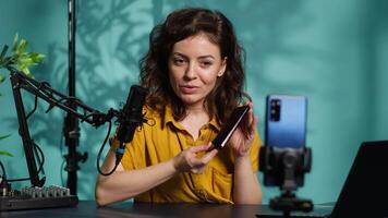 Smartphone on holder capturing footage of reviewer discussing with viewers about newly released cellphone. Influencer chats with fans during stream, filming latest generation phone review, camera A video