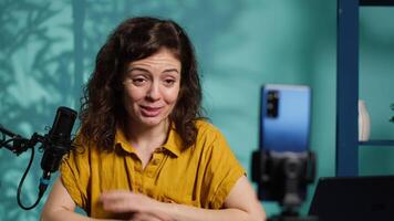 Smiling woman in studio using smartphone on phone holder to record , greeting subscribers. Happy online star hosts q and a session with viewers using cellphone, saluting fans, camera A video