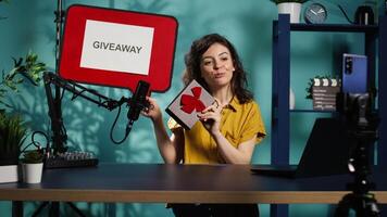 Smiling woman holding empty copy space sign, excited to do giveaway for subscribers. Media star happy to offer packed gift with ribbon to viewers, holding cardboard bubble speech cutout, camera B video