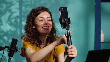Smiling woman in studio using smartphone on selfie stick to record , greeting subscribers. Joyous entertainer hosts q and a session with viewers using cellphone, saluting fans, camera A video
