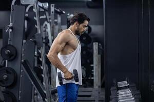 Muscular sport man is practice weight training on double dumbbells for biceps and triceps muscle inside gym with dark background for exercising and workout photo