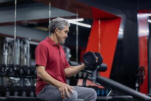Asian senior man wearing sportswear doing weight lifting on single dumbbell to gain bicep muscle while having exercise training in the gym for longevity and retirement concept photo