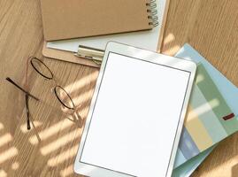 A workspace setup with a tablet, notebooks, glasses, and stationery on a wooden table illuminated by soft sunlight photo