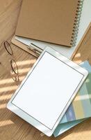 A workspace setup with a tablet, notebooks, glasses, and stationery on a wooden table illuminated by soft sunlight photo