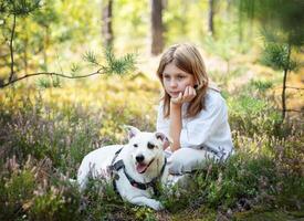 A young child sits thoughtfully beside a loyal dog in a serene forest surrounded by vibrant greenery and blooming flowers photo