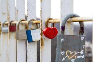 Close-up of various padlocks, including a red heart-shaped lock, attached to a rusty white metal railing symbolizing love, commitment, and memories on a bridge photo