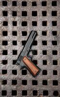 Black handgun with wooden grip resting on a rusted, industrial metal grate with square openings. A rugged, high-contrast shot perfect for illustrating security, danger, or weapon-related themes. photo