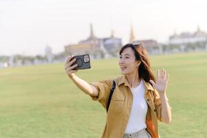 Traveler asian woman in her 30s making a livestream or selfie on smartphone while explores Wat Phra Kaew emerald Buddha. Share the wonders of Thai heritage through her journey. photo