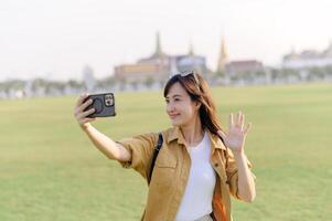 Traveler asian woman in her 30s making a livestream or selfie on smartphone while explores Wat Phra Kaew emerald Buddha. Share the wonders of Thai heritage through her journey. photo