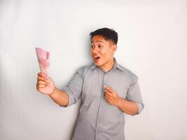 Young Asian pointing gesture his hand while holding banknotes and shows happy expression wearing grey shirt isolated on white background photo