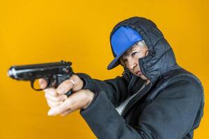 portrait of middle aged woman dressed as angry rapper in black tracksuit taking aim to shoot with gun posing over colorful background photo