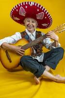 portrait of a happy middle aged woman smiling in front of a yellow background enjoying playing guitar dressed as a Mexican mariachi with sombrero photo