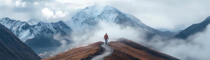 Hiker stands on a mountain trail with snow-capped peaks and clouds in the background, embracing the serene and majestic landscape. photo