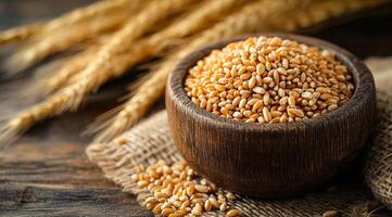 A rustic wooden bowl overflows with golden wheat grains, surrounded by dried wheat stalks and placed on textured burlap fabric. photo
