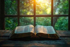 Open holy bible resting on a wooden table in front of window with sunlight shining through greenery photo