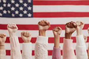 Hands with raised fists show solidarity and unity in front of a blurred American flag, symbolizing strength, activism, and diversity. photo