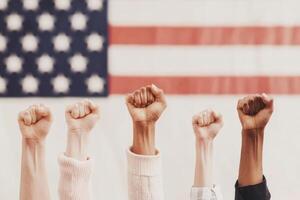 Hands with raised fists show solidarity and unity in front of a blurred American flag, symbolizing strength, activism, and diversity. photo