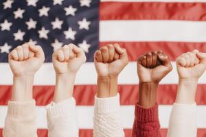 Hands with raised fists show solidarity and unity in front of a blurred American flag, symbolizing strength, activism, and diversity. photo