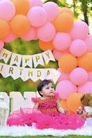 A baby girl in a pink dress sitting in front of a birthday arch photo