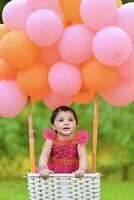 A baby girl sitting in a basket with pink and orange balloons photo