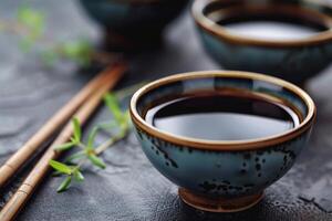 Bowls filled with soy sauce resting on dark surface with chopsticks photo
