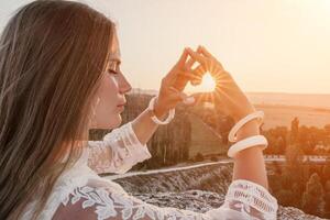 Happy woman in white boho dress making heart sign with hands on sunset in mountains. Romantic woman with long hair standing with her back on the sunset in nature in summer with open hands. photo