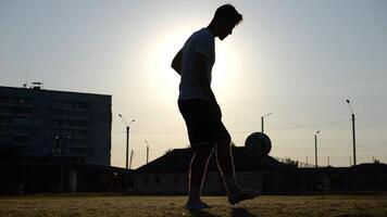 Professional footballer juggling soccer ball on stadium at sunset. Young man kicking ball at green field. Sportsman practicing tricks at meadow with sunlight at background. Freestyle football concept photo