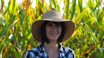 Happy smiling female farmer looks into camera standing near corn field. Portrait of adult beautiful agronomist in straw hat with maize meadow at background. Agricultural business concept. Close up photo