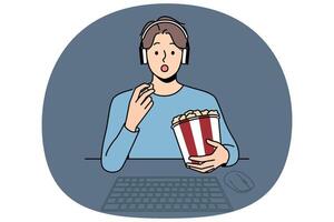 Man with popcorn watches movie sitting at table with computer and looks at screen intrigued vector