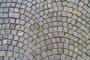 A charming cobblestone street showcases a radial pattern of gray stones, lined with colorful buildings. Weeds peek through the cracks, adding a touch of nature to this historic scene, ideal for tourists. photo