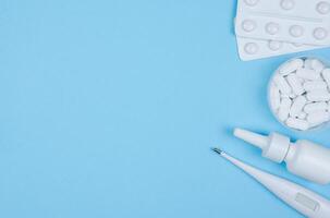Blisters with tablets, a thermometer, and nasal spray on blue background. Flat lay with copy space. Stock photo