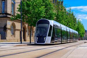 Luxembourg, A modern silver tram moves swiftly along the street, creating a motion blur, while a vintage stone building with many windows stands alongside, surrounded by trees and urban scenery. photo