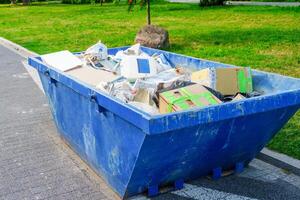 A blue dumpster is sitting in the park. The dumpster is filled with various types of garbage including cardboard boxes, paper, and plastic bottles. The dumpster is sitting on a paved path next to the grass photo