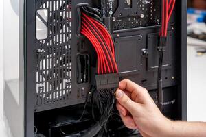 A hand connects a black ATX power supply cable to an Asus motherboard inside a sleek black case. The motherboard features red accents on a black PCB, with a tempered glass side panel for visibility. photo