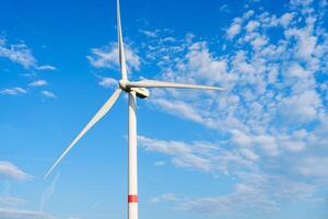 A lone wind turbine stands tall against a blue sky with fluffy clouds. The turbines white blades spin slowly, capturing wind energy and converting it into clean electricity. photo