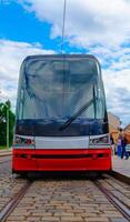 The tram is sleek and modern, featuring a large windshield and a vibrant red body. It glides along a brick track, surrounded by blurred buildings, people walking by, and a lively urban atmosphere. photo
