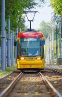 The image features a modern yellow and red tram on a bustling streetof Warsaw. The tram is sharply in focus against a blurred background, emphasizing its vibrant colors and creating a dynamic sense of motion. photo
