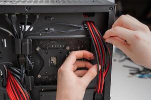A black and red themed ATX mid-tower PC build with a tempered glass side panel and ARGB lighting. A hand holds the back panel open, showcasing the motherboard, CPU cooler, graphics card, and RAM. photo