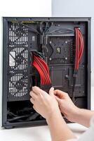 A person is carefully installing computer hardware components inside a sleek black case. The case features a glass side panel, showcasing the colorful red cables and the power supply unit visible inside. photo
