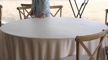 Event planner smooths out the wrinkles on a beige tablecloth covering a round table with wooden chairs video