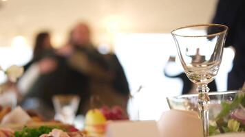 An empty wine glass sits on a table with a blurred image of newlyweds receiving congratulations in the background video