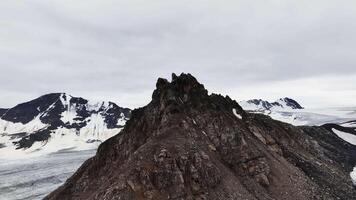 Aerial view of mountains in summer. Drone top view of high cliffs, snow, sky with clouds. Mountain peaks. Mystical landscape, smooth drone flight video