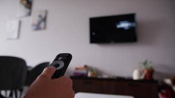 Close-up of a woman's hand holding a TV remote control and changing TV channels. In the background of the hand is a wall with a TV in out of focus. Blurred TV video