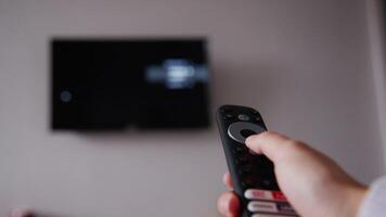 Close-up of a woman's hand holding a TV remote control and changing TV channels. In the background of the hand is a wall with a TV in out of focus. Blurred TV video
