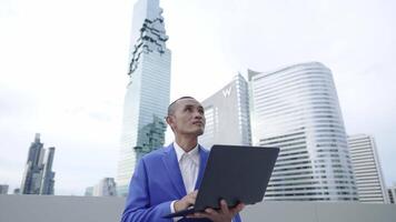 A businessman in a blue suit stands outdoors in front of skyscrapers, holding a laptop and looking upward. The image conveys innovation, technology, and ambition in an urban corporate setting. video