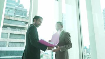 A businessman holding folders approaches a colleague who is standing by a large window, gazing out at the city skyline. The scene conveys reflection and corporate planning. video
