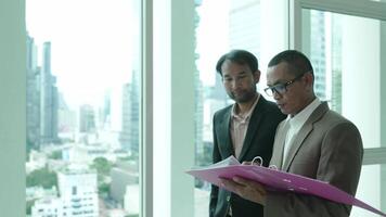 Two business professionals stand by a large window in an office, reviewing documents in a folder. The cityscape in the background reflects a corporate and urban environment. video